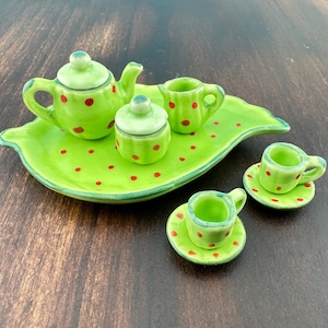 Puede incluir: Un juego de té de cerámica en miniatura de color verde brillante con lunares rojos. El juego incluye una tetera, azucarero, lechera y dos tazas de té con platillos, todo dispuesto sobre una bandeja con forma de hoja. Un artículo decorativo encantador.