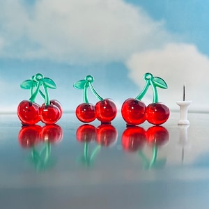 Puede incluir: Tres pares de chinchetas rojas con forma de cereza con tallos y hojas verdes, y una sola chincheta blanca. Las chinchetas están dispuestas sobre una superficie reflectante contra un fondo de cielo azul con nubes. Las cerezas son translúcidas.