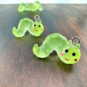 May include: Three translucent green caterpillar charms with silver-colored loops for hanging. Each caterpillar has a smiling face with black eyes, pink cheeks, and a red mouth. The charms are on a dark wood surface.