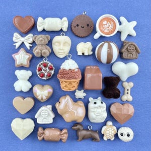 Könnte beinhalten: Eine Sammlung von Miniatur-Anhängern in verschiedenen Formen und Farben, darunter Herzen, Bären und eine Kaffeetasse. Die Anhänger sind in Braun-, Weiß- und Cremetönen gehalten und vor einem blauen Hintergrund platziert. Die Sammlung umfasst eine Vielzahl von Texturen und Designs.