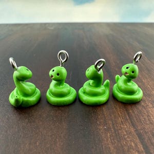 May include: Four small, bright green snake charms with silver-colored loops for hanging. Each snake has a coiled body, a small head with black eyes, and a smiling mouth. The charms are displayed on a dark brown surface.