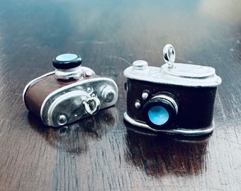 May include: Two miniature silver camera charms with brown and silver details. One camera charm has a blue lens and the other has a silver lens.