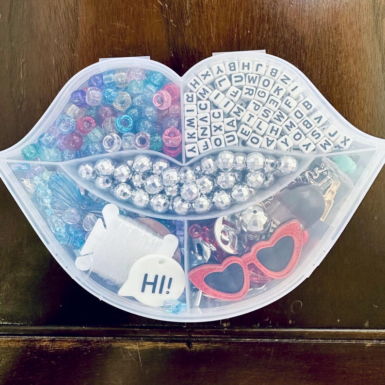 May include: A clear, lip-shaped container filled with various craft supplies. The container is divided into sections holding beads, letter beads, silver beads, red heart-shaped sunglasses, and a white tag with "HI!" written on it.