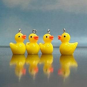May include: Four yellow rubber duck charms with orange beaks and silver jump rings. The ducks are arranged in a row on a reflective surface.