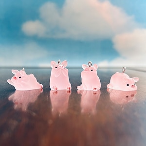May include: Four pink pig charms with silver loops. The pigs are in different poses, including sitting, lying down, and looking up.