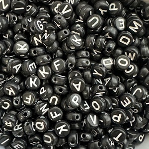May include: A close-up image of a pile of black letter beads. The beads are round and have white letters on them. The letters are all different and include letters like A, B, C, D, E, F, G, H, I, J, K, L, M, N, O, P, Q, R, S, T, U, V, W, X, Y, and Z.