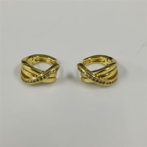 以下が含まれることがあります： クロスデザインの2つのゴールドカラーのイヤーカフ。各カフには、小さな透明な宝石の列があります。イヤリングは白い表面に表示され、その複雑なディテールとエレガントなスタイルを示しています。