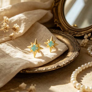 Puede incluir: Un par de pendientes de botón dorados con forma de estrella con una piedra central azul claro. Los pendientes se exhiben en una bandeja plateada de estilo vintage con un paño blanco. Un collar de perlas y un espejo decorativo están en el fondo.