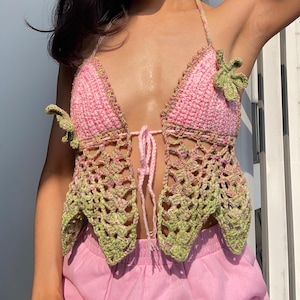 Puede incluir: Top halter de ganchillo rosa y verde con cierre de lazo y adornos de hojas. El diseño calado es visible. El esquema de color y el estilo sugieren una estética bohemia y artesanal.
