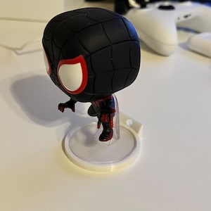 Puede incluir: Figura de vinilo Funko Pop de Miles Morales Spider-Man, negra y roja. La figura tiene ojos blancos grandes con contornos rojos y está en posición agachada. Está montada sobre un soporte de plástico transparente.