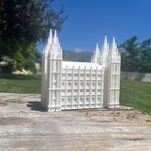 May include: A white architectural model of a building with multiple spires and a rectangular body. The model is placed on a weathered wooden surface, with a green lawn and blue sky in the background. The model is a detailed replica.