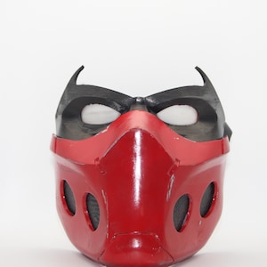 May include: A red and black mask with a glossy finish. The mask features a red lower section with three oval openings on each side and a black upper section with eye openings and pointed tips.