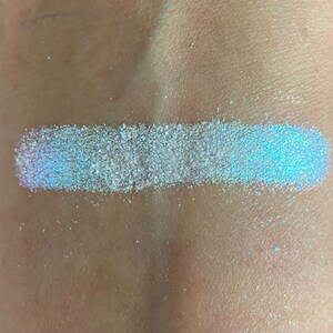 May include: A swatch of iridescent blue glitter on a person's skin. The glitter is a light blue color with a shimmery finish.