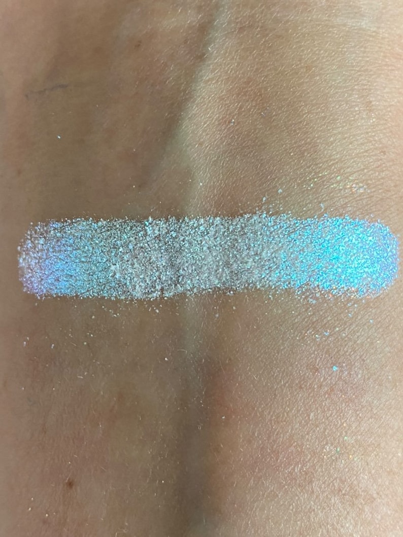 May include: A swatch of iridescent blue glitter on a person's skin. The glitter is a light blue color with a shimmery finish.
