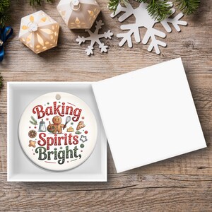May include: A round, white Christmas ornament with the text "Baking Spirits Bright" in red and green, featuring gingerbread man and baking-themed illustrations. The ornament is in a white gift box, with a wooden background.