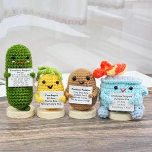 May include: Four crocheted amigurumi toys, each representing a different food item: a pickle, an apple, a potato, and a dumpster fire. Each toy is holding a sign with a positive message. The pickle says "Emotional Support Pickle", the apple says "Fine Apple", the potato says "Positive Potato", and the dumpster fire says "Emotional Support Dumpster Fire".