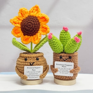 May include: Two crocheted emotional support plants in brown pots with happy faces. The sunflower plant has green leaves and a yellow and brown flower. The cactus plant has green stems with pink flowers. Both plants have a white card with the text "Emotional Support Plant" and a message about life being tough but remembering that you are too.
