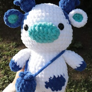 May include: A handmade plush cow toy in white and blue. The cow has a blue nose, ears, and spots, with a matching blue bag hanging from a blue strap. The toy has black eyes and is made of soft, textured yarn.