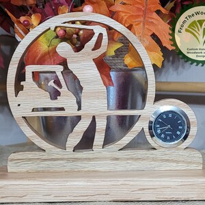 May include: A wooden golf themed clock with a golfer silhouette, golf cart, and clock face. The clock is on a wooden base and has the text "FromTheWoodpile.com Custom Handcrafted Woodwork and Vinyl Designs" on the side.