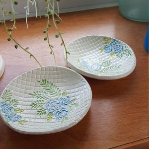 Könnte beinhalten: Zwei weiße Keramikteller mit einem gewebten Muster und blauen und grünen Blumendesigns.