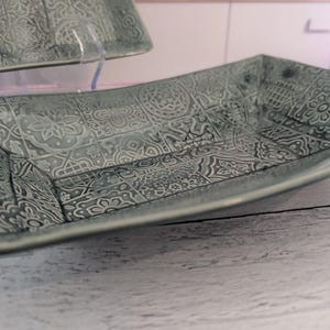 Pode incluir: Dois pratos retangulares de cerâmica com um esmalte verde sálvia e um padrão em relevo repetitivo de intrincados motivos geométricos e florais. Os pratos têm bordas ligeiramente curvas e um acabamento brilhante com um design de azulejos.