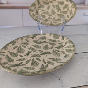 Op de afbeelding: Twee ovale keramische borden met een crèmekleurige achtergrond. De borden zijn versierd met een patroon van groene libellen en varenbladeren. De borden worden gepresenteerd op een transparante plastic standaard.