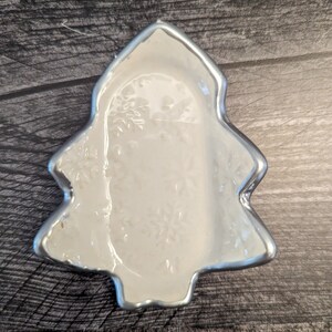 Puede incluir: Un plato de cerámica blanca con forma de árbol de Navidad con un borde plateado. El plato tiene un diseño de copo de nieve.