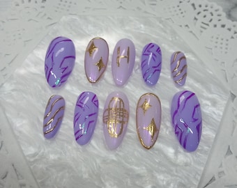 Pode incluir: Um conjunto de dez unhas artificiais em vários tons de roxo e lavanda. Cada unha apresenta detalhes dourados, incluindo formas geométricas e linhas. As unhas estão dispostas sobre uma superfície branca texturizada.