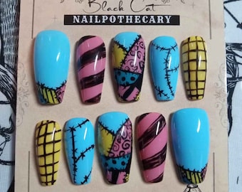Puede incluir: Un conjunto de diez uñas postizas personalizadas con varios diseños. Las uñas presentan una base azul claro con costuras negras, rayas rosas y marrones y patrones de patchwork. El texto "The Black Cat Nailpothecary" está en la parte superior.