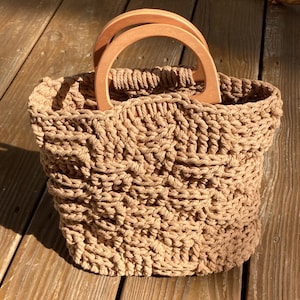 May include: A brown crocheted tote bag with wooden handles. The bag is made of a thick, textured yarn and has a rectangular shape.