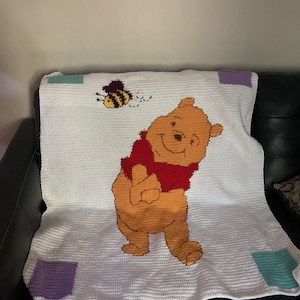 Puede incluir: Una manta de ganchillo blanca con un gráfico central de un oso amarillo con una camiseta roja y una pequeña abeja volando encima. La manta tiene un fondo blanco con cuatro cuadrados en las esquinas en colores lila claro y verde azulado.