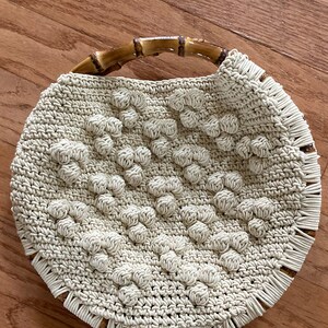May include: A round, crocheted bag with a woven texture and a brown bamboo handle. The bag has a fringe trim around the bottom.