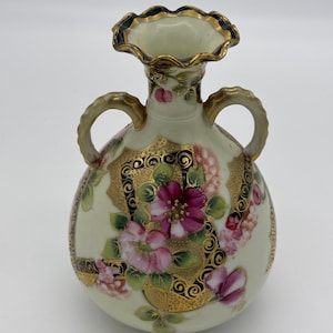May include: A small, cream-colored porcelain vase with two handles. The vase is decorated with pink and purple flowers, gold accents, and black swirling designs.