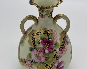 May include: A small, cream-coloured porcelain vase with two handles. The vase is decorated with pink and purple flowers, gold accents, and black swirling designs.