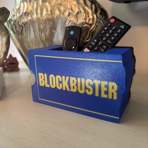 May include: A blue and yellow Blockbuster-themed remote control holder. The holder has the Blockbuster logo in yellow on a blue background. Two remotes are inside the holder, one black and one small black remote.