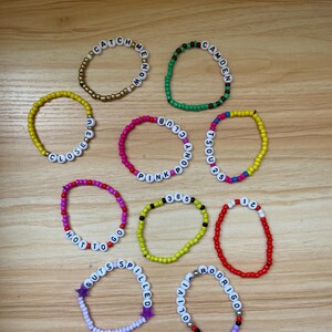 Może przedstawiać: Dziesięć kolorowych bransoletek z koralików z różnymi słowami i frazami napisanymi na nich. Bransoletki są wykonane z małych, okrągłych koralików w różnych kolorach, w tym żółtym, zielonym, różowym, czerwonym i fioletowym. Słowa na bransoletkach to "Catch Me", "Close 2 U", "Hot To Go", "Guts Spilled", "Olivia Rodrigo", "Camden", "Pink Pony Club", "365", "110" i "TSOU".