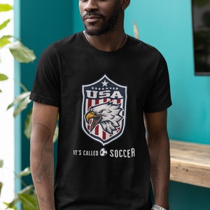 May include: Black t-shirt with a graphic of a bald eagle inside a shield with the text "USA" and stars. Below the shield is the text "It's called soccer" with a soccer ball graphic.