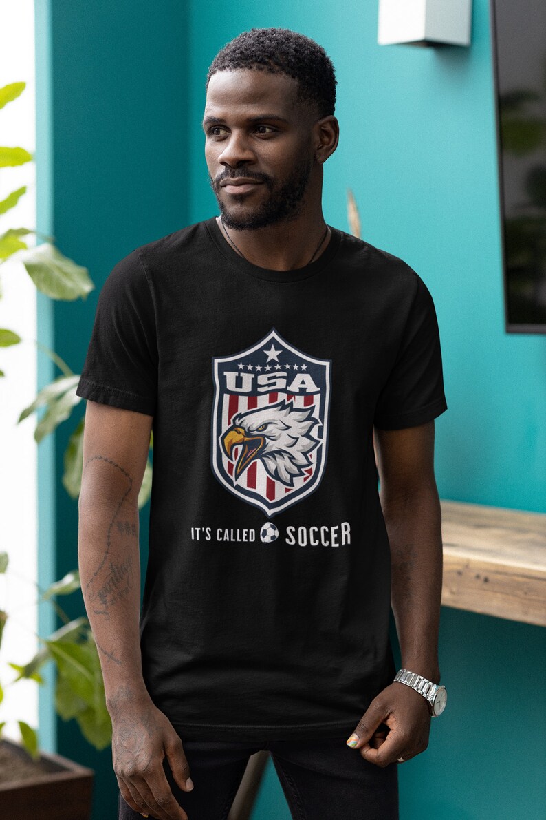 May include: Black t-shirt with a graphic of a bald eagle inside a shield with the text "USA" and stars. Below the shield is the text "It's called soccer" with a soccer ball graphic.