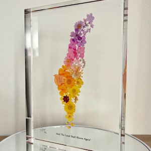 May include: A rectangular clear acrylic block containing pressed flowers arranged in a vertical gradient from yellow at the bottom to purple at the top. The text 'And The Land Shall Bloom Again' is printed in black at the bottom of the block.
