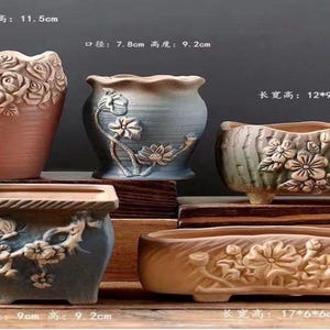 May include: Five ceramic planters with embossed floral designs. Top left: pink rose pot, 8cm diameter, 11.5cm height. Top center: blue lotus pot, 7.8cm diameter, 9.2cm height. Top right: green floral trough, 12x9x7.2cm. Bottom left: blue square pot, 9cm height. Bottom right: beige floral trough, 17x6x6cm.