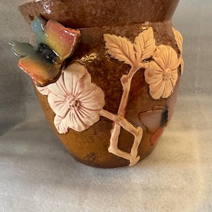 May include: A brown ceramic planter with a raised floral design. The planter features a butterfly, leaves, and flowers in shades of brown, pink, and white.