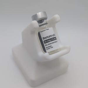 Könnte beinhalten: Ein weißer, handförmiger Halter hält ein kleines Glasfläschchen mit einem silbernen Verschluss. Das Fläschchen hat ein Etikett mit dem Text "Retatrutik 10 mg / 3 mL For Subcutaneous Multiple Dose Vial" und "Custom Text". Der Halter steht auf einem weißen Sockel.