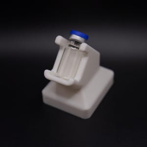 May include: A white plastic stand holding a small, clear glass vial with a blue cap. The stand has a rectangular base and a cradle to secure the vial. The background is a solid black.