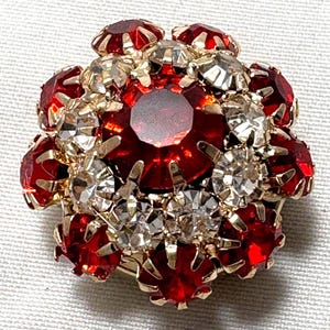 May include: A vintage floral brooch with a gold-tone setting. The brooch features a central large red gemstone surrounded by smaller red and clear rhinestones. The design is symmetrical and detailed, with a classic, elegant style.