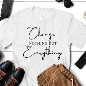 May include: White t-shirt with black text that reads "Change Nothing But Everything". The shirt is laid flat on a white wooden surface with a brown leather bag, a pair of black shoes, brown trousers, a pair of sunglasses, and a mobile phone.
