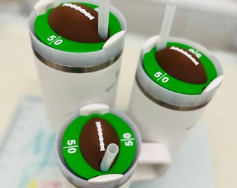 May include: Three white insulated tumblers with football-themed lids. Each lid features a brown football on a green field with the 50-yard line markings. White straws are inserted into the lids. The tumblers are designed for cold beverages.