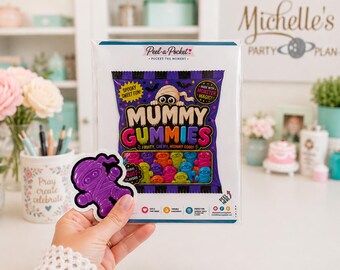May include: A clear plastic package of Mummy Gummies candy featuring a purple bag with a mummy illustration and colorful gummy figures. A separate purple mummy-shaped sticker is held in front of the package, which displays the text Peel-a-Pocket, Mummy Gummies, and Fruity, Chewy, Mummy Good.