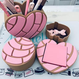 May include: A collection of pink and tan desk organizers shaped like concha pastries. The set includes a heart-shaped pen holder filled with pens and scissors, a small container with bows, and a round container with a heart-shaped lid holding a pink sticky note pad.