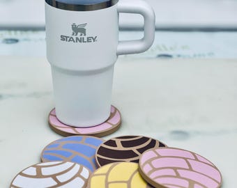 May include: A white Stanley tumbler with a pink and brown lid and straw, and a set of colorful, round coasters. The coasters feature a concha bread design in various colors, including white, blue, yellow, and pink.