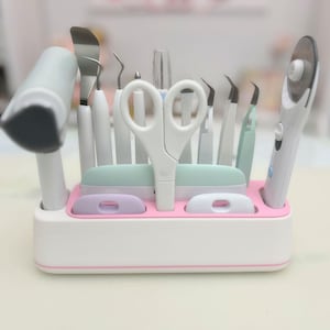 May include: A white and pink organizer holding various craft tools, including scissors, a rotary cutter, and several other tools with white handles. The organizer has a pink base and a white exterior with a pink trim.